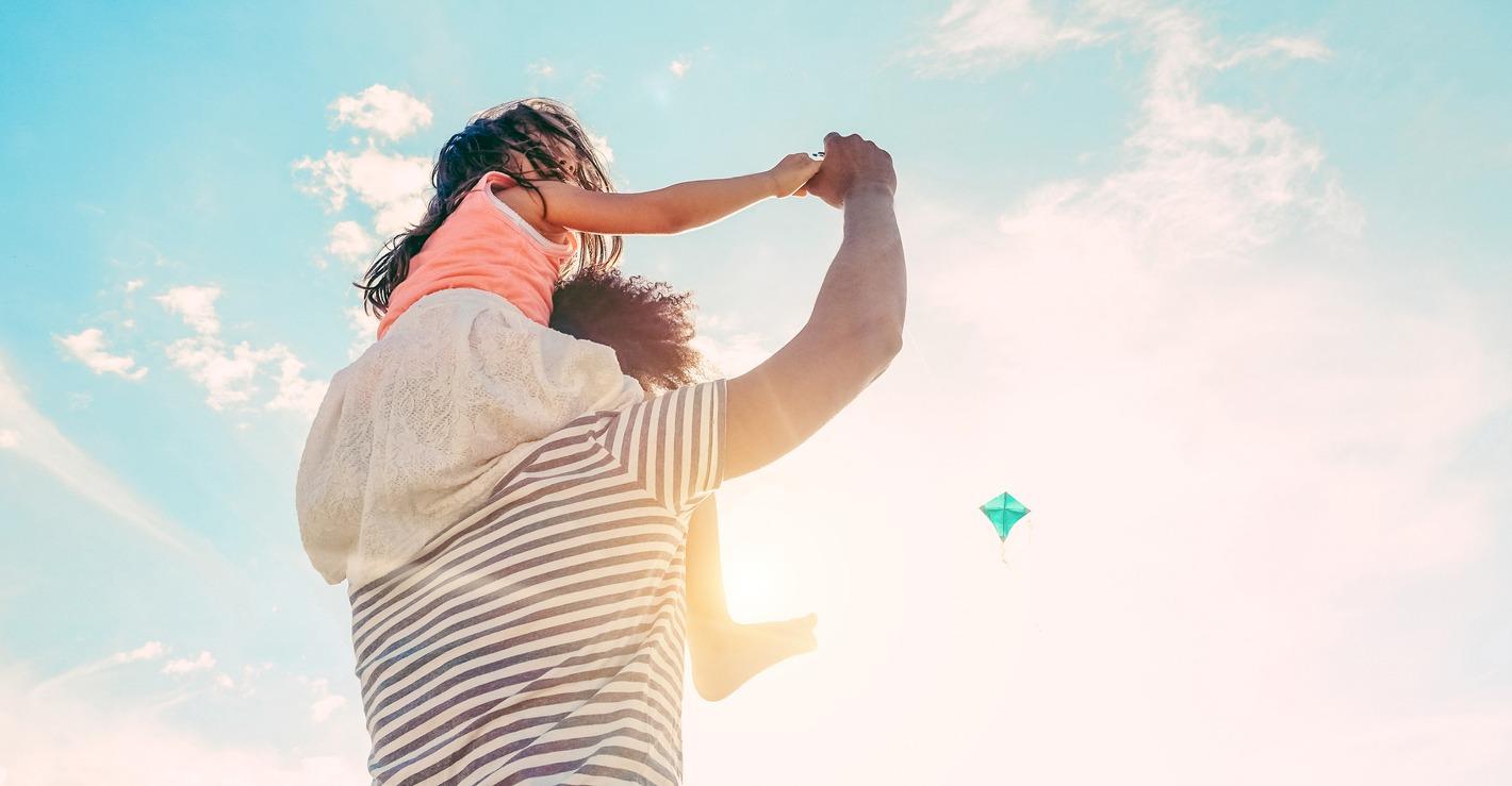 Father and Daughter playing on the beach while watching someone fly a kite.